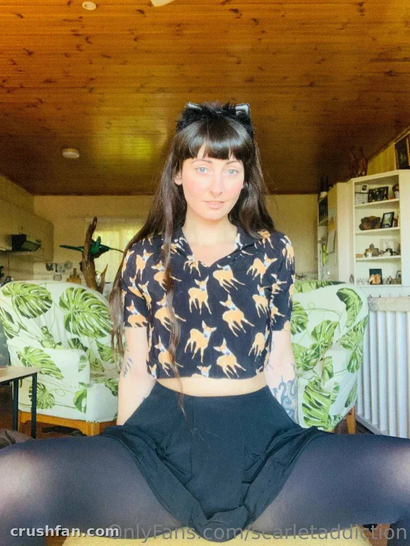 Scarlet in a leaf-print crop top and black skirt seated between palm-print armchairs indoors