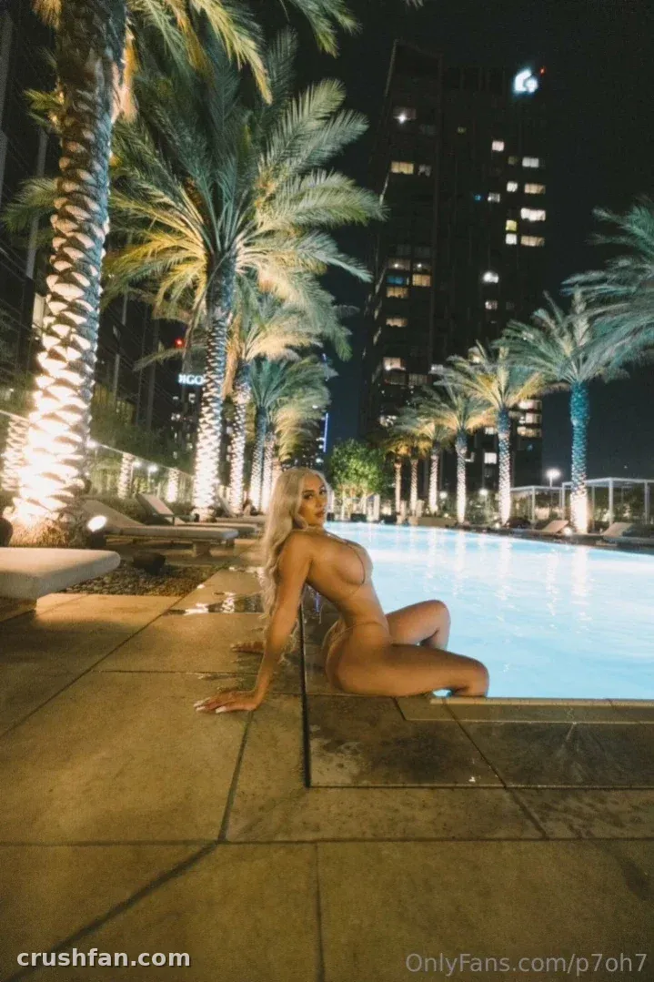 Pamela en bikini sentada junto a una piscina iluminada por la noche con palmeras y edificios de la ciudad detrás de ella.