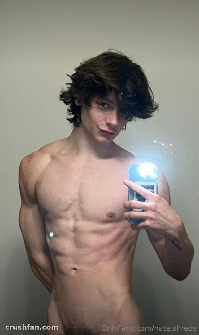 Hombre sin camiseta con cabello oscuro ondulado tomándose una selfie de espejo con una cámara con flash contra una pared lisa.