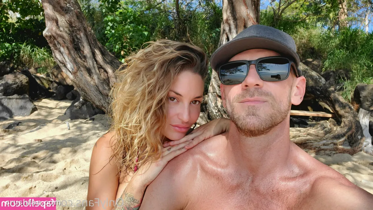 Kissa Sins posando con un acompañante en una playa soleada frente a los árboles