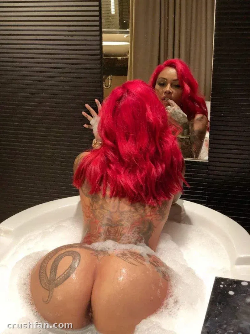 Brittanya com cabelo rosa sentada em um banho de espuma, vista de costas com o reflexo de um espelho.
