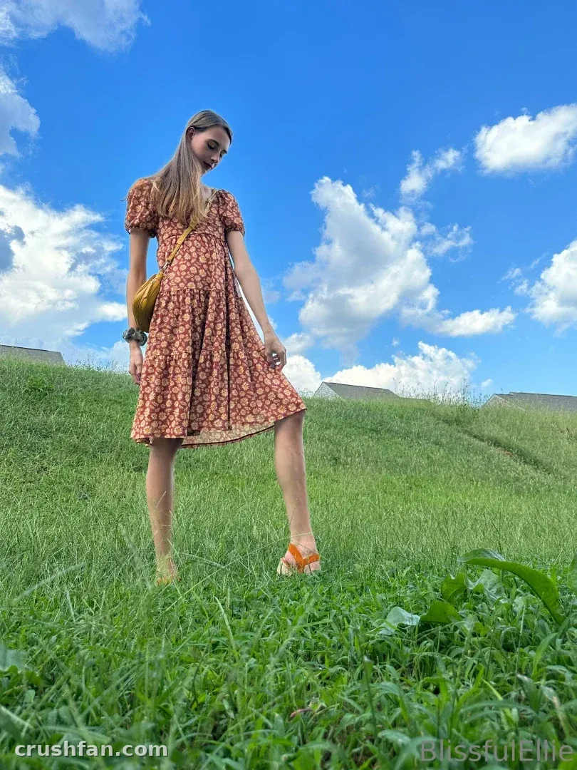 Ellie em um vestido floral em pé em uma colina gramada sob um céu azul