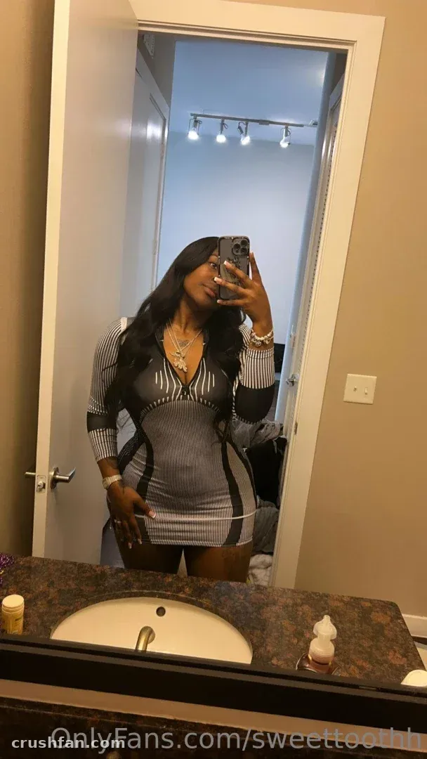 Sweettoothh con un vestido gris a rayas tomándose una selfie en el espejo de un baño.