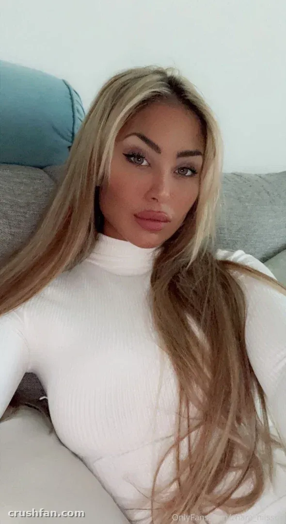 Zhara Nilsson con un suéter de cuello alto blanco sentada en un sofá gris en interiores