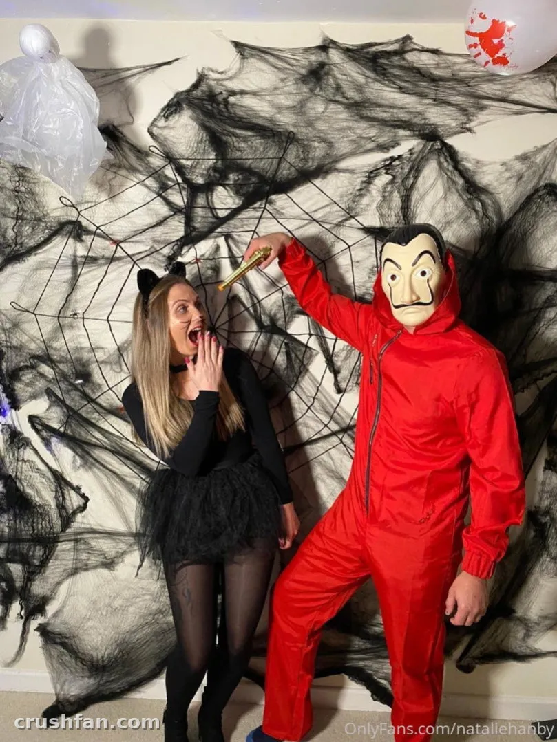 Frau in einem schwarzen Katzenkostüm neben einer Figur in einem roten Overall und Maske vor künstlichen Spinnweben