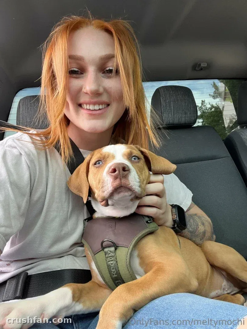Mulher sorridente de cabelo ruivo segurando um cachorro no banco de trás de um carro, vestindo uma camisa branca.