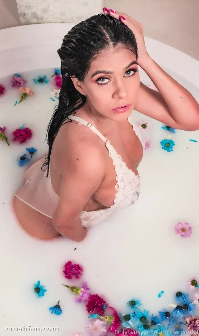 Carolina Romo em um banho leitoso com flores rosas e azuis flutuantes, usando um bralette de renda branca.