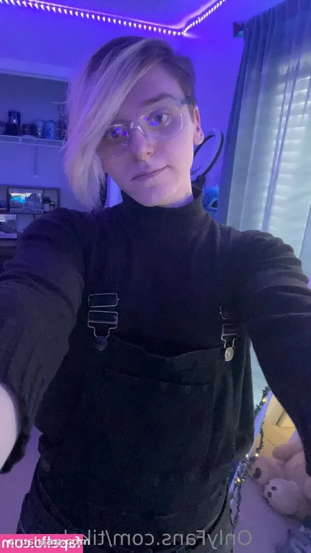 Tika in a black turtleneck and overalls taking a selfie in a purple-lit room.