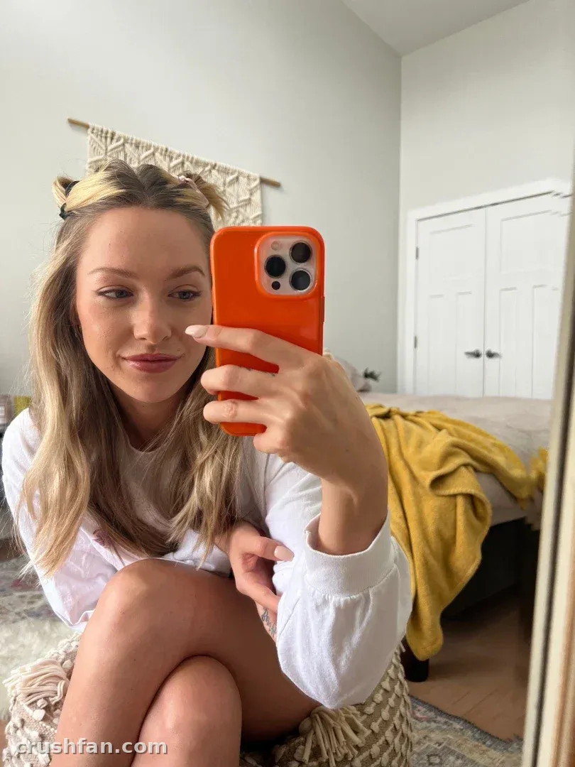 Forrest in a white top taking a mirror selfie with an orange phone case in a bright room.