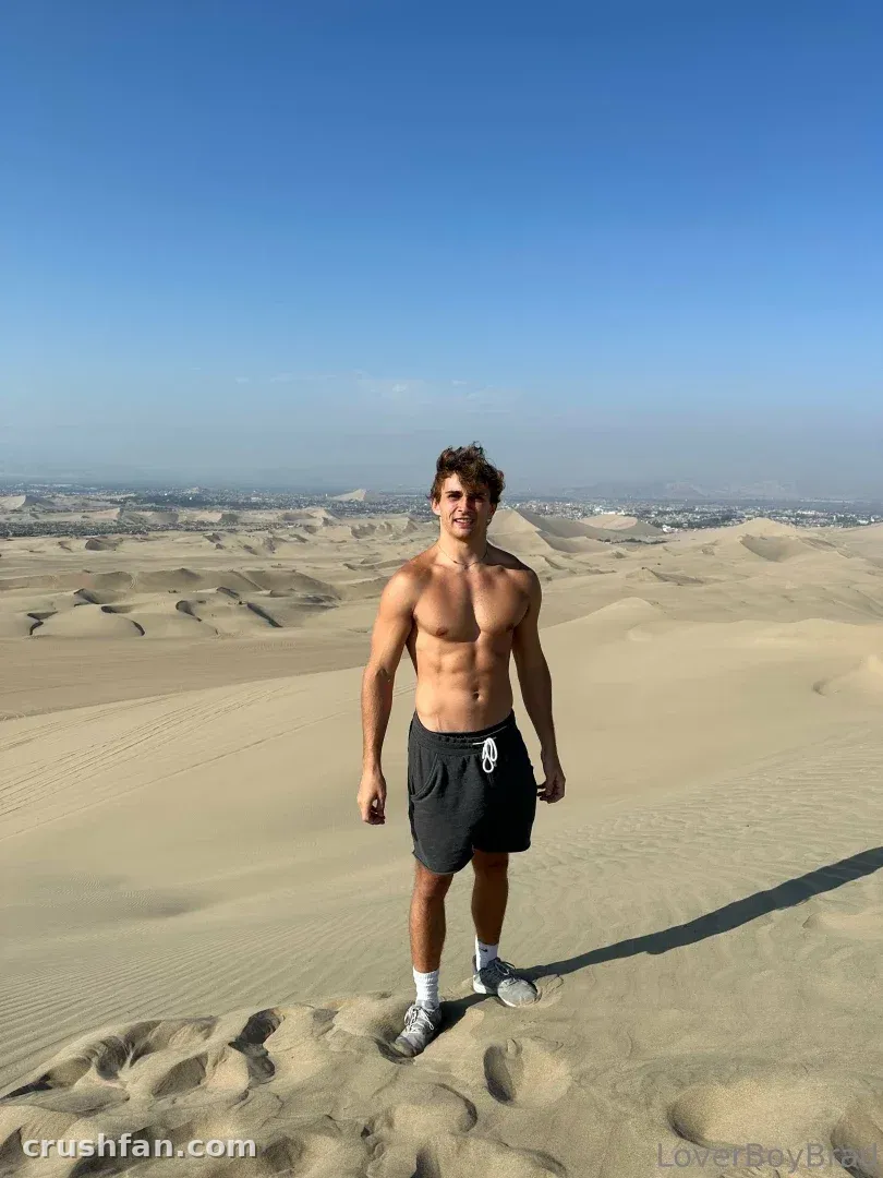 Leo shirtless in dark shorts standing on desert sand dunes under a clear blue sky