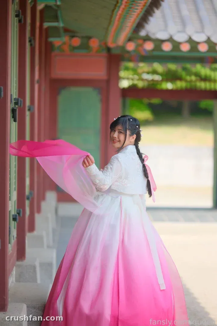 quqco dans un hanbok rose et blanc tenant un tissu fluide à côté de piliers rouges