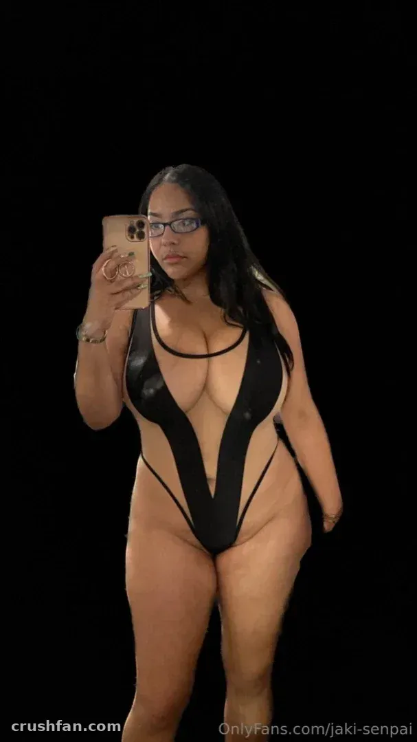 Criadora com um body preto brilhante recortado tirando uma selfie no espelho contra um fundo escuro