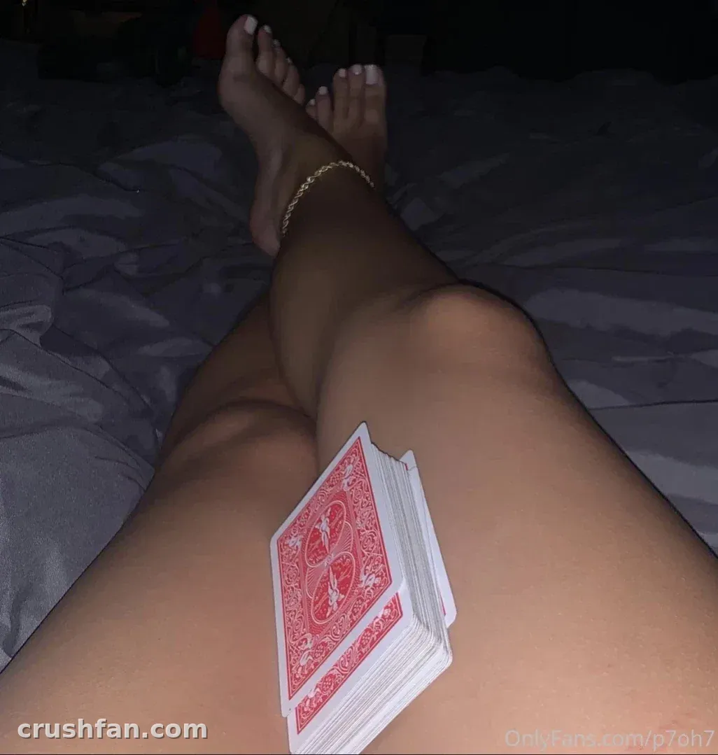 Pamela reclining on a bed with a deck of cards resting on her leg in dim light.