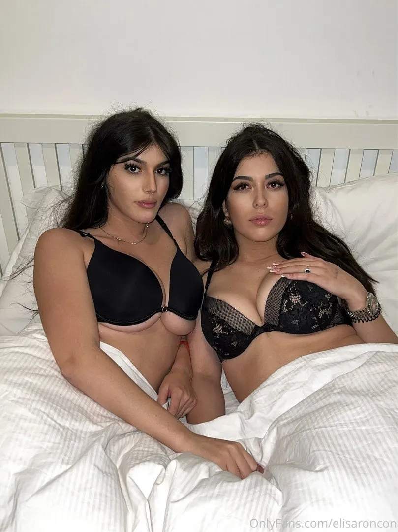Two women in black lingerie sitting together on a white bed with striped sheets