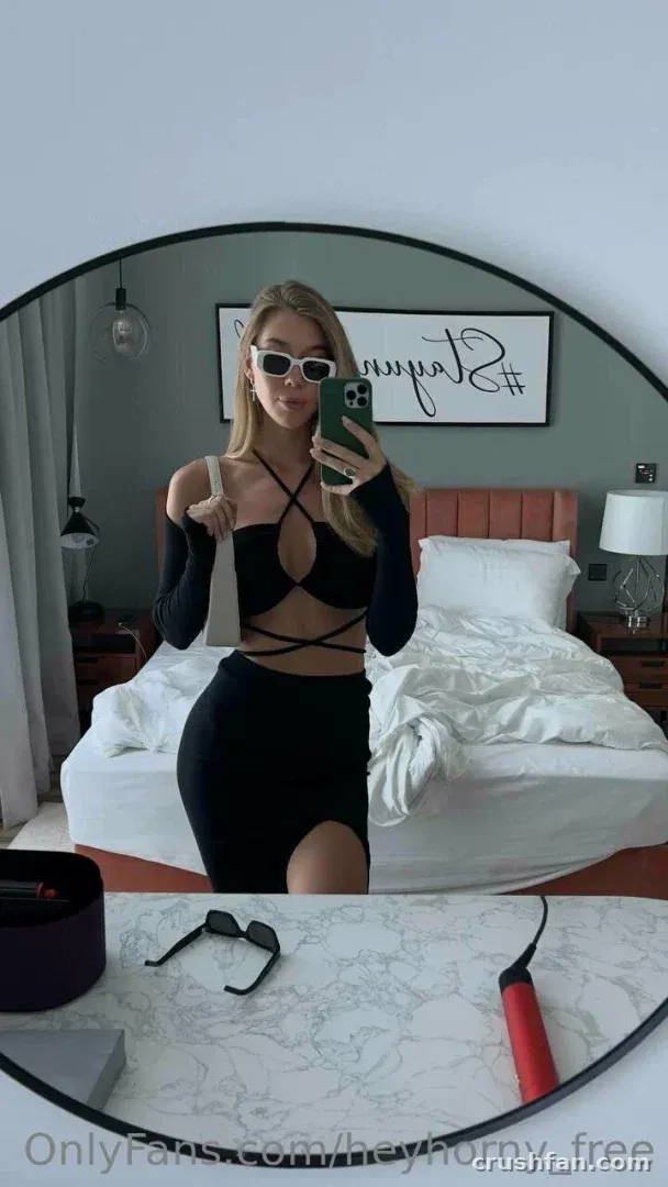 HeyHorny com uma blusa preta com recortes e saia tirando uma selfie no espelho em um quarto.