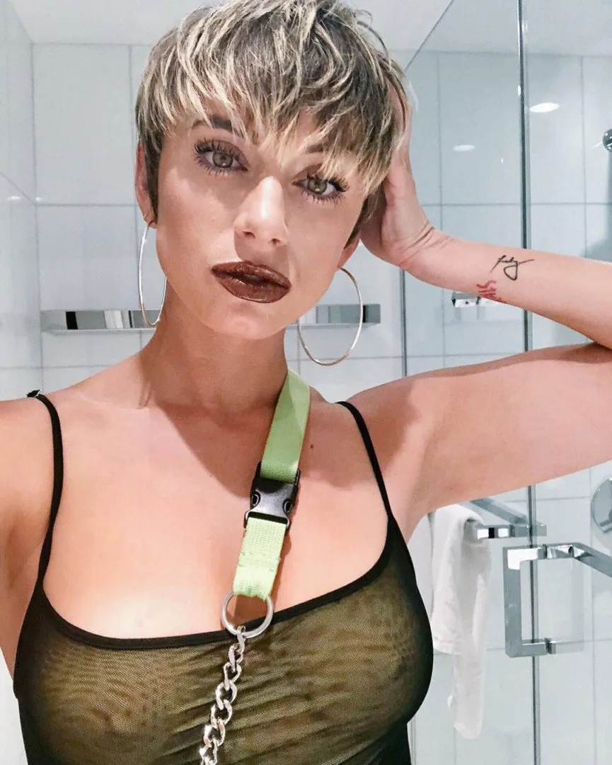 Yesjulz in a black mesh top with a green strap and large hoop earrings in a tiled bathroom.