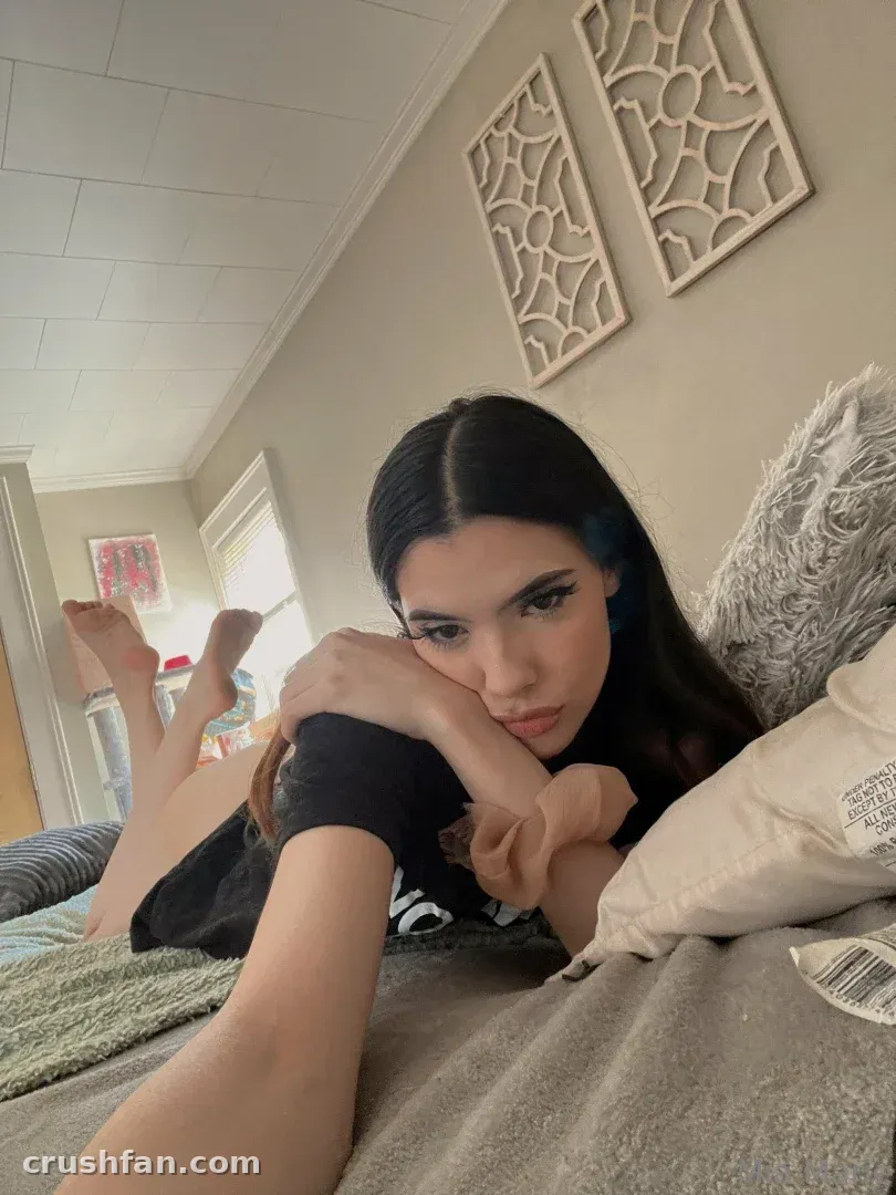 Mia Marie lying on a grey bed in a black top, looking at the camera in a softly lit room.