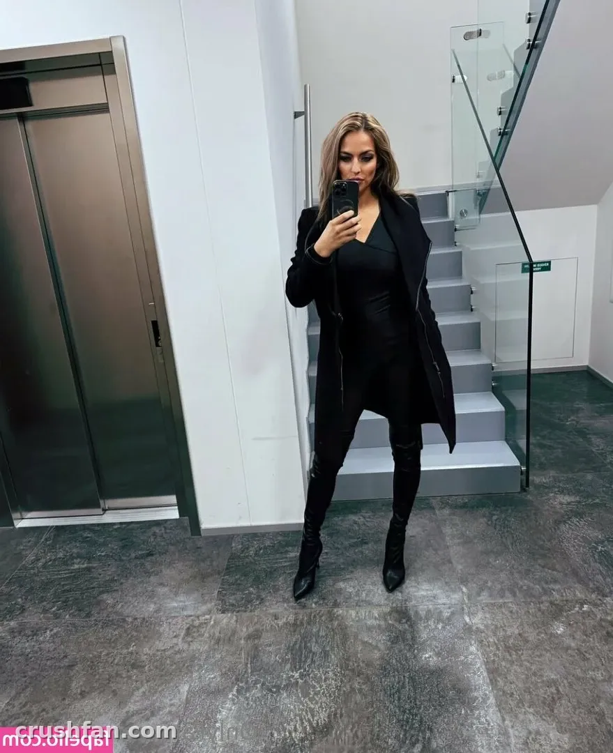 Creator in an all-black coat and boots taking a phone selfie in a modern stairwell