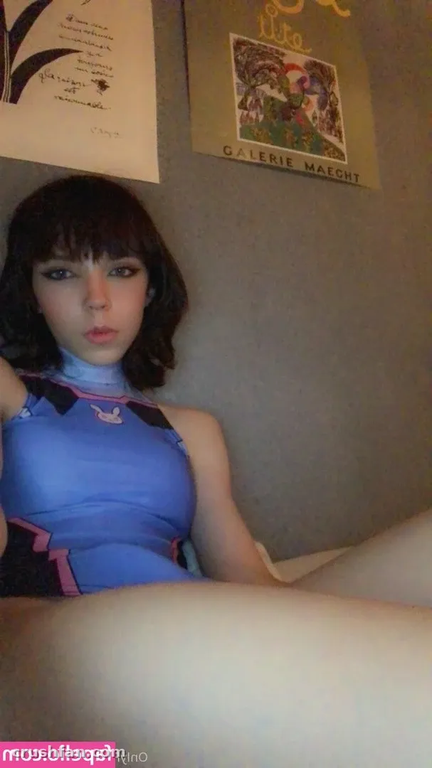 Remi in a blue cosplay top posing indoors in front of a grey wall with two posters.