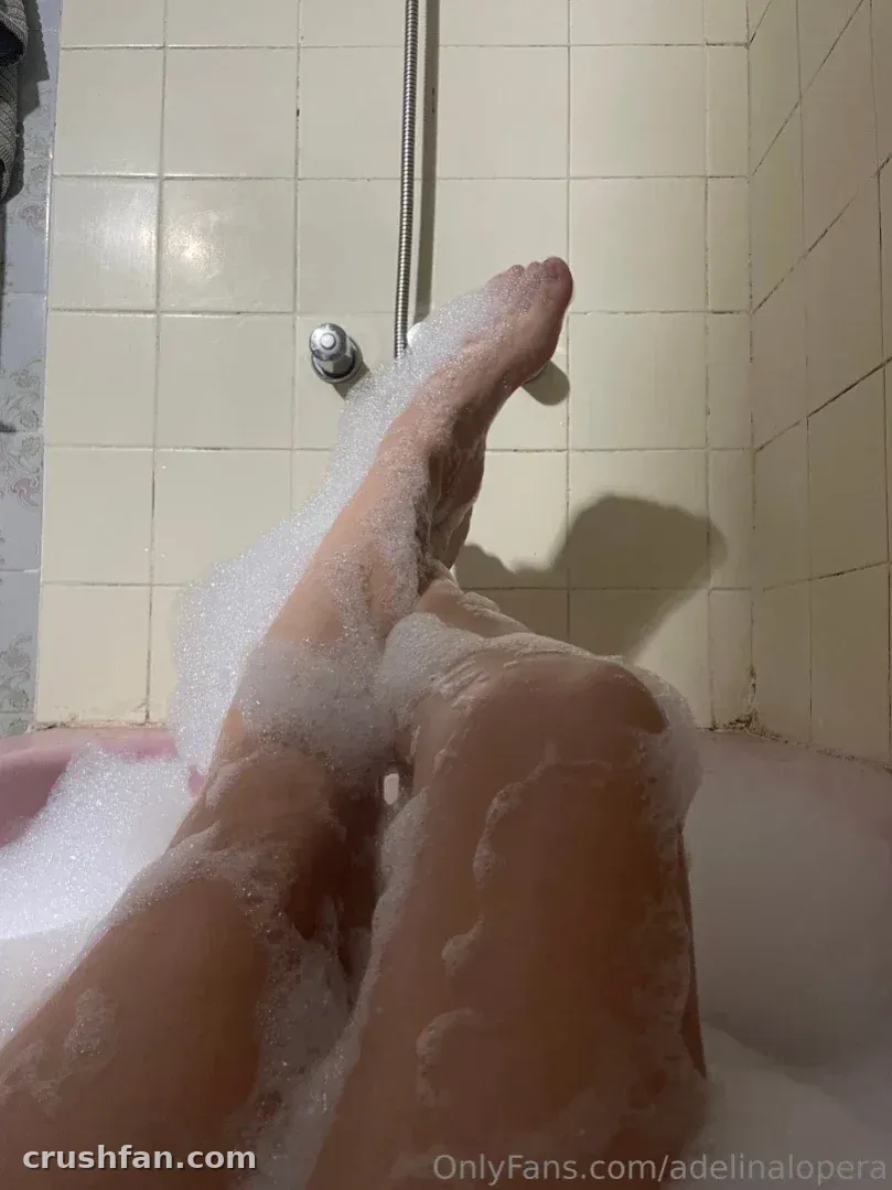 Legs raised above a foamy bathtub against cream bathroom tiles