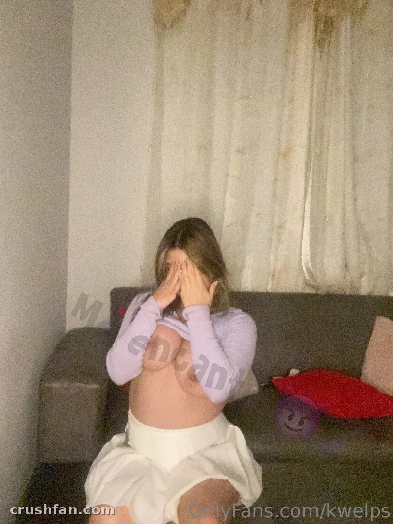 Kwelp seated on a grey sofa with hands over her face, wearing a lilac top and white skirt.