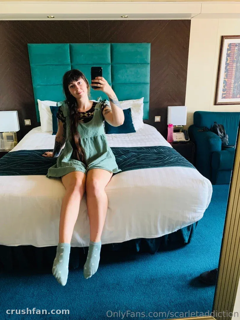 Scarlet con un romper verde tomándose una selfie frente al espejo mientras está sentada en una cama de hotel