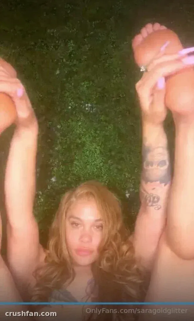 Sara Gold deitada na grama com os braços levantados, mostrando uma tatuagem no antebraço e unhas rosas.