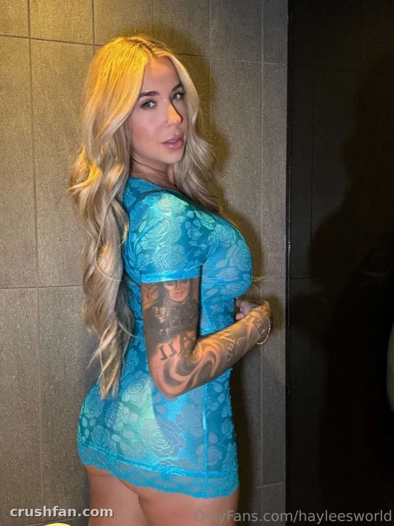 Criadora em um vestido de renda azul posando ao lado de uma parede de azulejos cinzas em ambiente interno