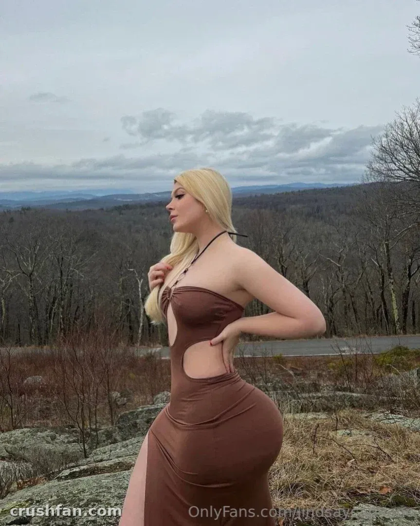Lindsay Capuano in a brown cutout dress standing on a rocky overlook under a cloudy sky.