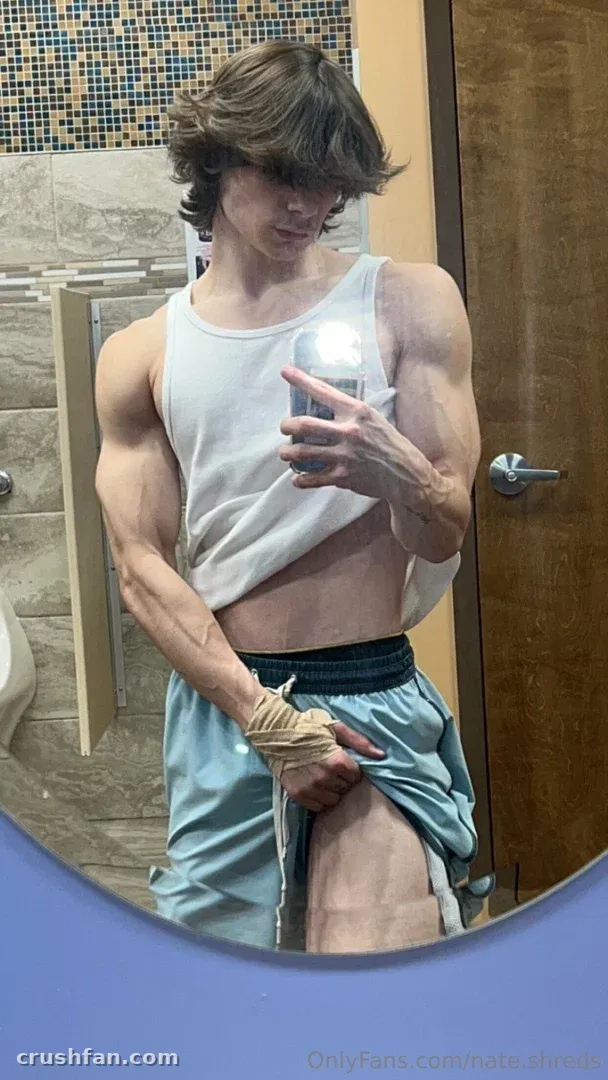 Nathan in a white tank top and blue shorts taking a mirror selfie in a tiled bathroom.