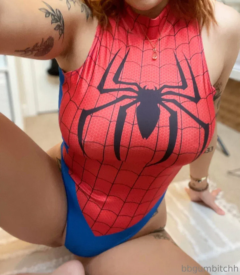 Feya Quinn seated indoors wearing a red and blue spider print bodysuit with tattoos visible.