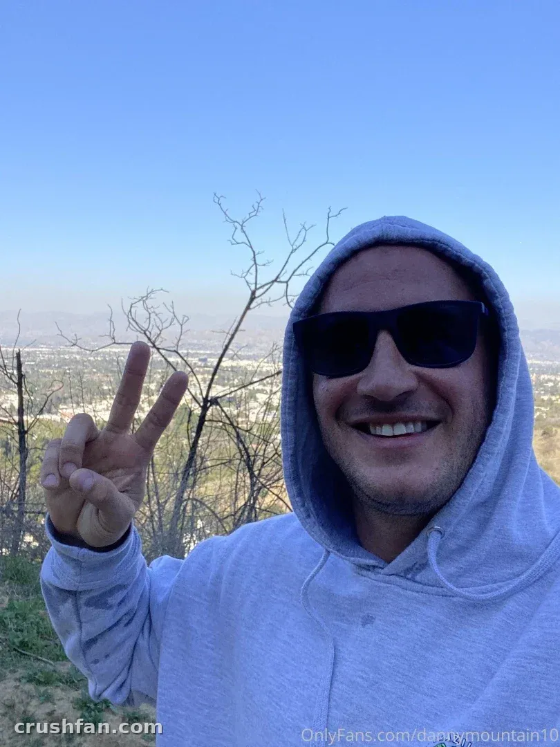 Danny Mountain en sweat à capuche gris et lunettes de soleil faisant un signe de paix en extérieur