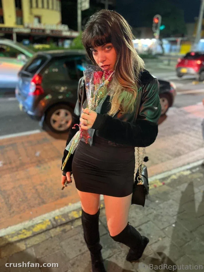 Chechu in a black leather jacket and skirt holding a bouquet on a night street