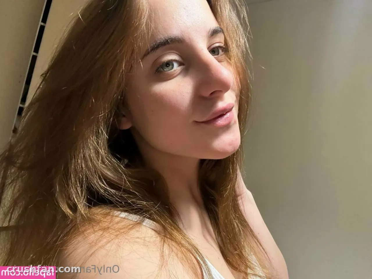 Selfie de ambiente interno em close-up de uma mulher com cabelos longos e ondulados usando uma blusa branca