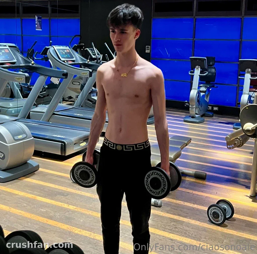 Alex está de pie sin camiseta en un gimnasio sosteniendo mancuernas, con cintas de correr y ventanas iluminadas en azul detrás de él.