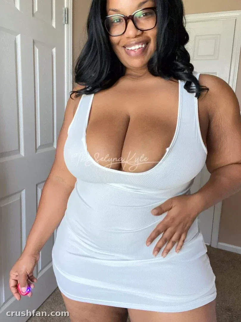 Mulher sorridente com vestido branco e óculos em pé em ambientes internos junto a portas brancas