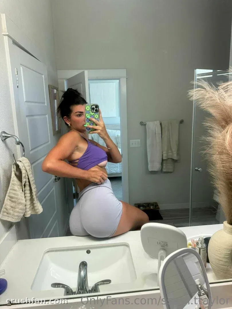 Sam Taylor taking a mirror selfie in a bathroom wearing a purple top and grey shorts.