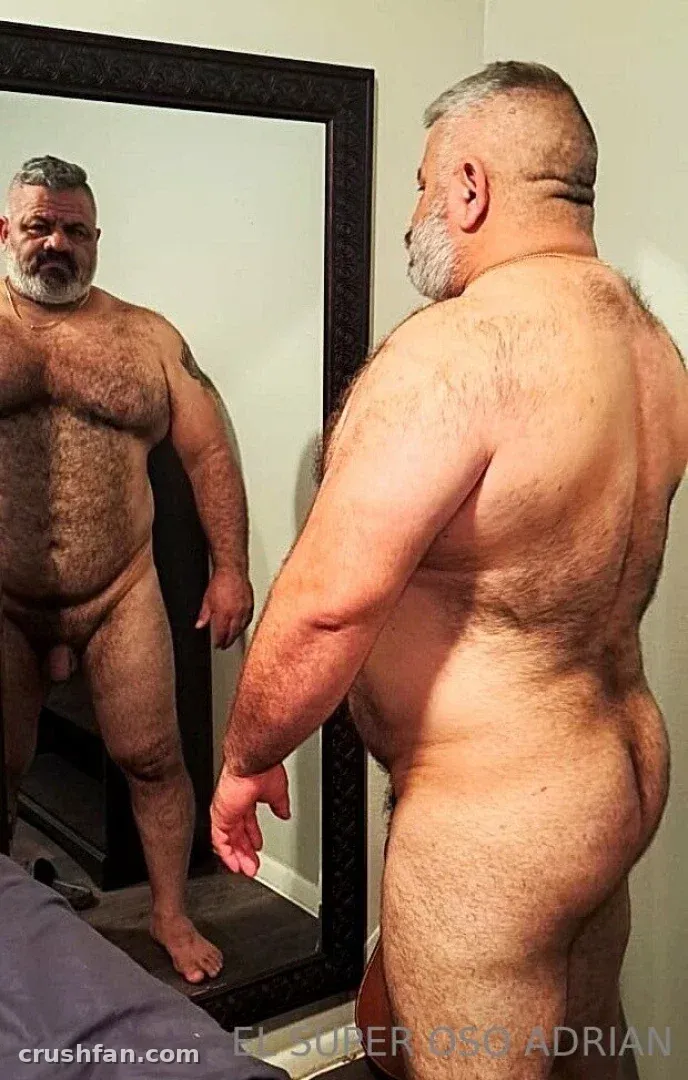 EL SÚPER OSO ADRIAN standing beside a tall framed mirror in a softly lit room