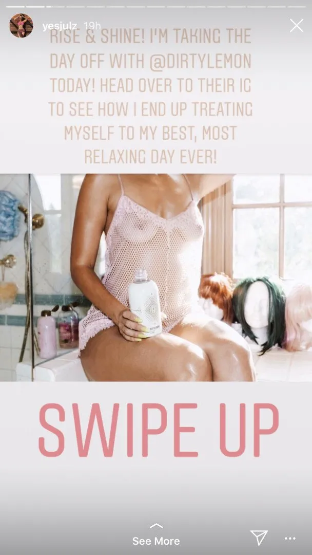 Yesjulz sitting on a bathtub edge in a sheer white bodysuit holding a bottle.