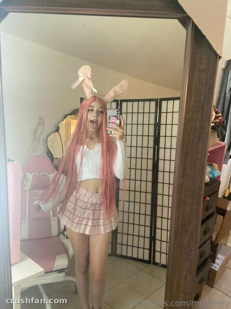 Moxie Rey com peruca rosa e orelhas de coelho tirando uma selfie no espelho em uma sala com uma cadeira rosa.
