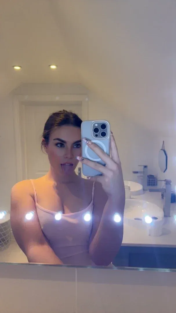 Lauren Alexis takes a mirror selfie in a bathroom, wearing a pink top and holding a blue phone.