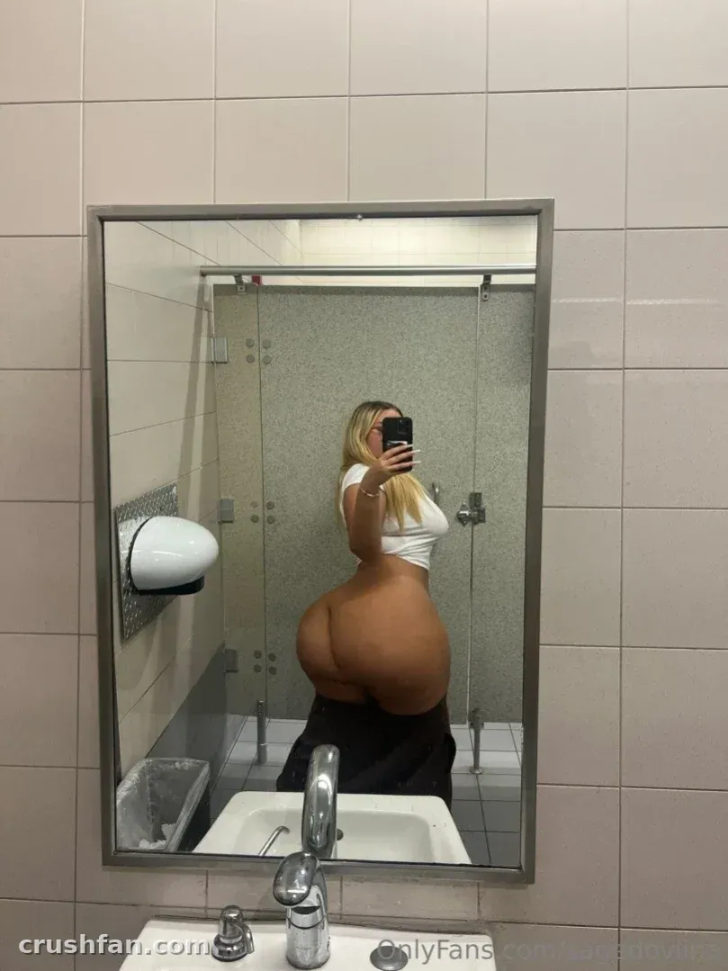 Sage Dovina en haut blanc prenant un selfie dans le miroir dans une salle de bain carrelée avec un lavabo devant.