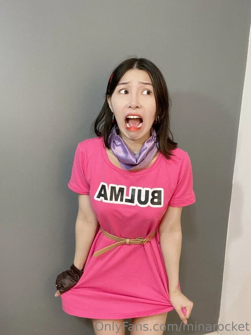 Mina Berry con un vestido rosa de eslogan y un pañuelo lila, de pie contra una pared gris.