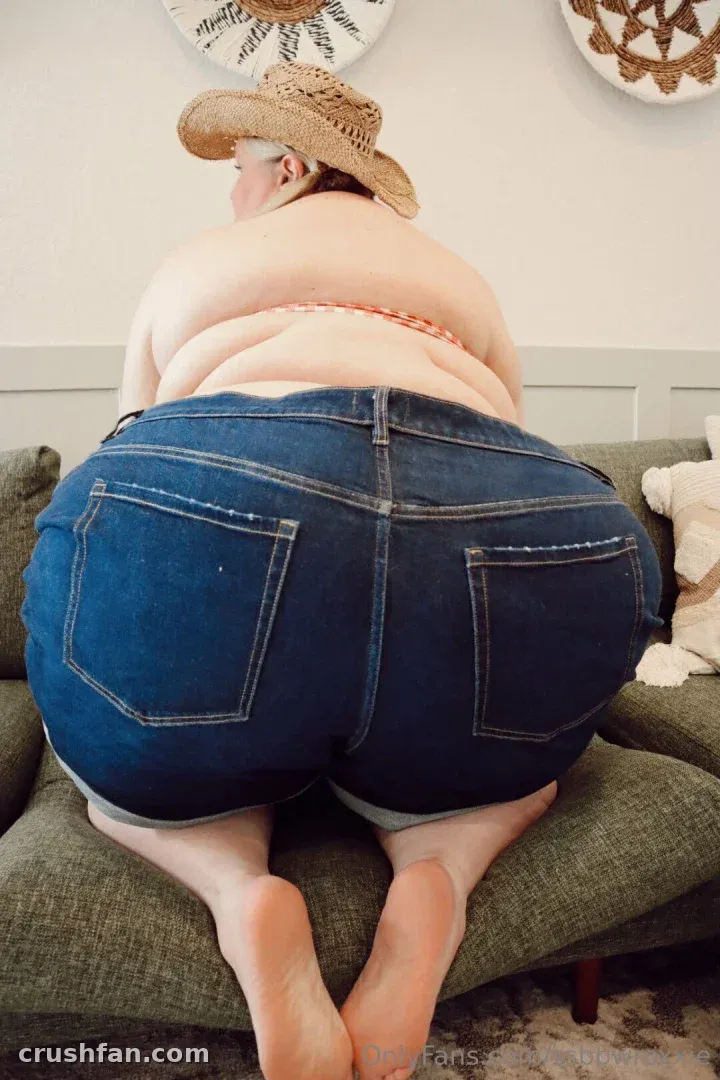 Supersized Roxxie kneeling on a green sofa in denim shorts and a straw hat.