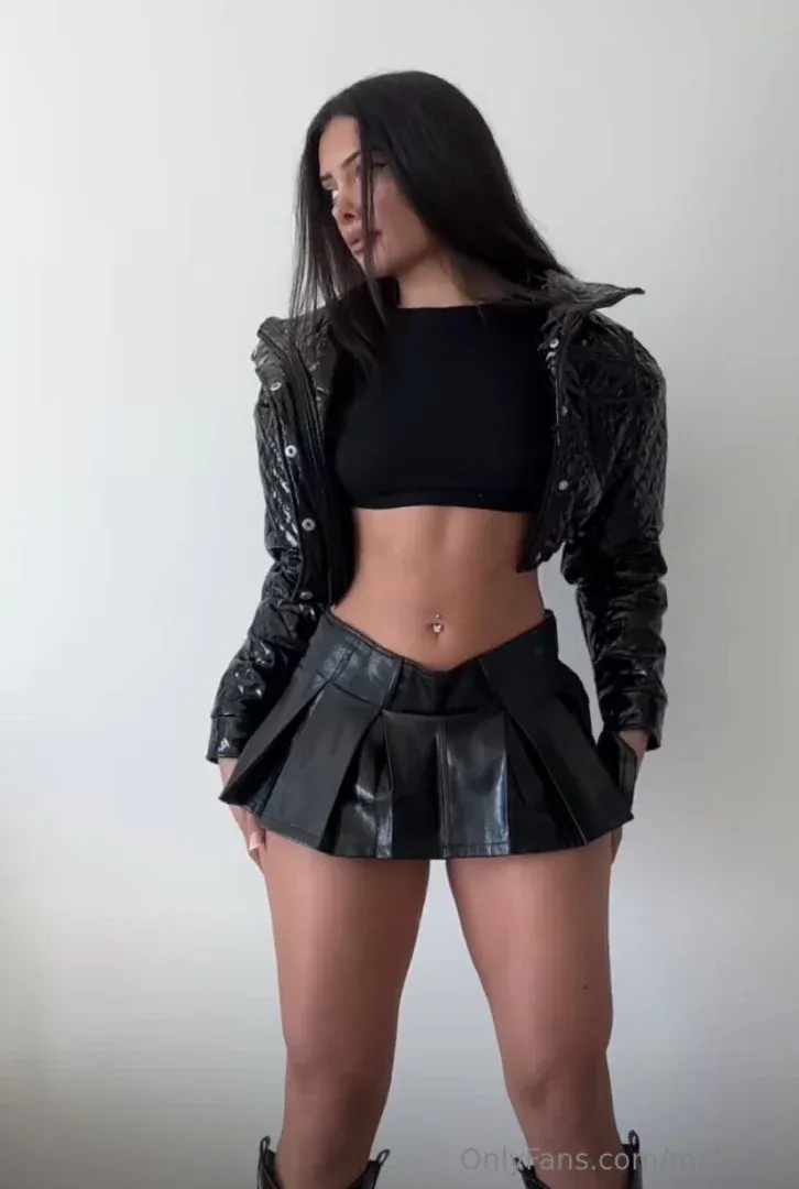 Melissa com uma jaqueta de couro preta e saia plissada contra uma parede lisa.