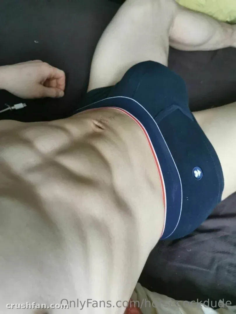 Le gars de Horsecock allongé sur un lit sombre en sous-vêtement bleu foncé avec une ceinture à rayures rouges.