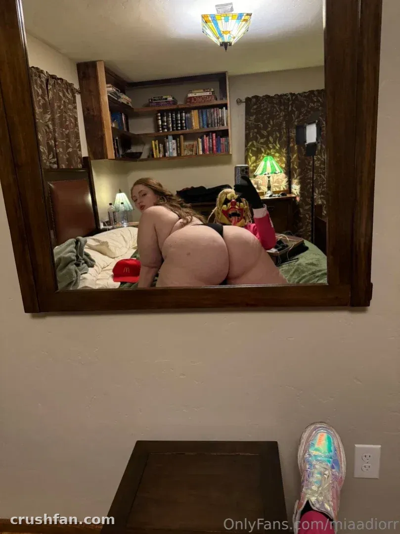 Reflet dans le miroir d'une femme posant sur un lit dans une chambre chaleureusement éclairée avec une étagère.