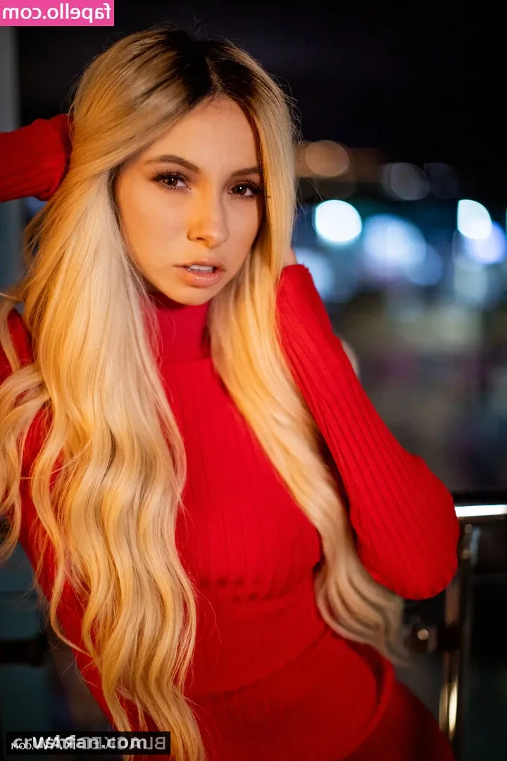 Mujer de largo cabello rubio ondulado con un cuello alto rojo posando ante las luces desenfocadas de una ciudad nocturna
