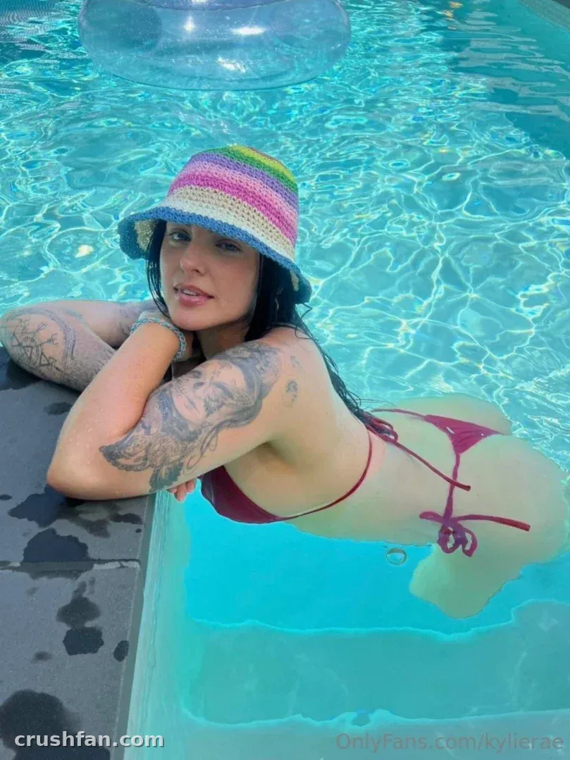 Kylie Rae in a striped bucket hat and magenta bikini leaning on a pool edge with turquoise water