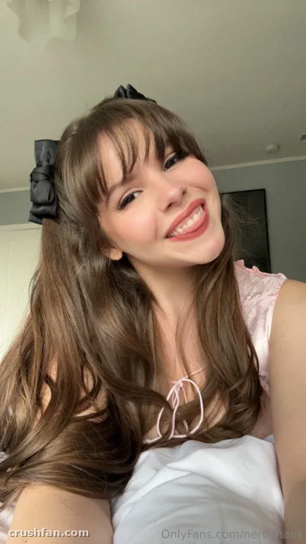 Hannah sorrindo em uma selfie próxima em ambiente interno, usando uma blusa rosa-claro e um laço preto no cabelo.
